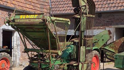 Cette photo permet de visualiser le bac à feuilles à l’arrière du tracteur,  le tapis élévateur de betteraves sur le flanc gauche de l’engin  et la trémie à betteraves positionnée au-dessus du pont avant du tracteur.